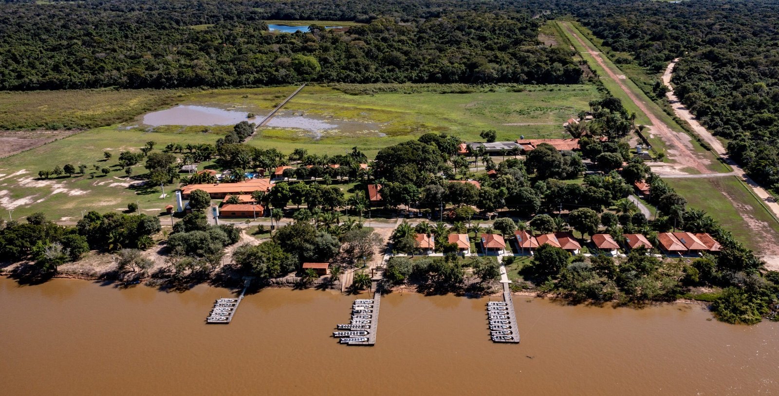 Aerial view of Hotel Porto Jofre Pantanal Norte on the Cuiabá River, with three piers lined with boats, red-roofed buildings among trees, a pool and an airstrip beside the forest