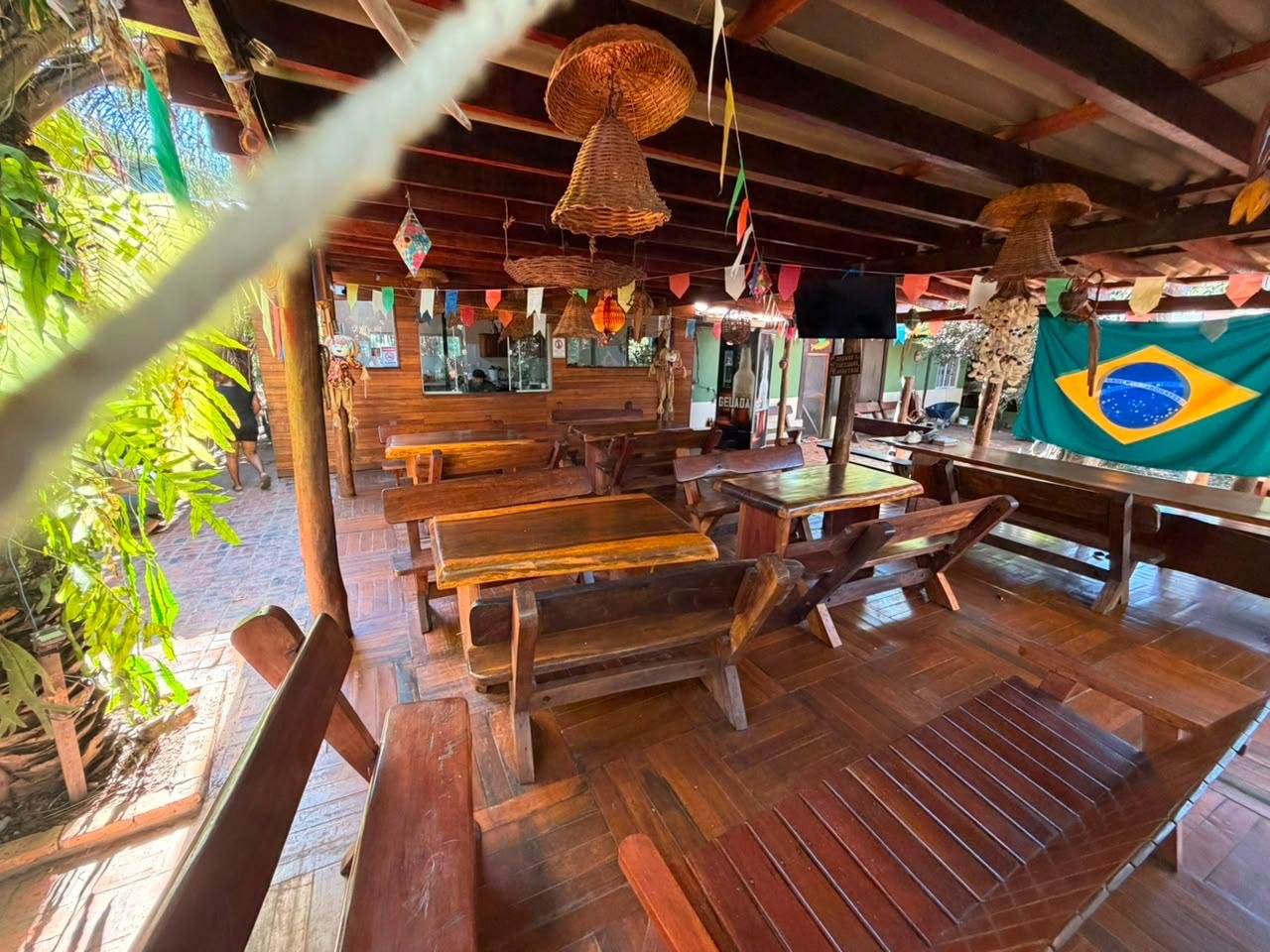 Covered seating area with rustic wooden tables and benches, woven pendant lamps, colorful bunting and a Brazilian flag