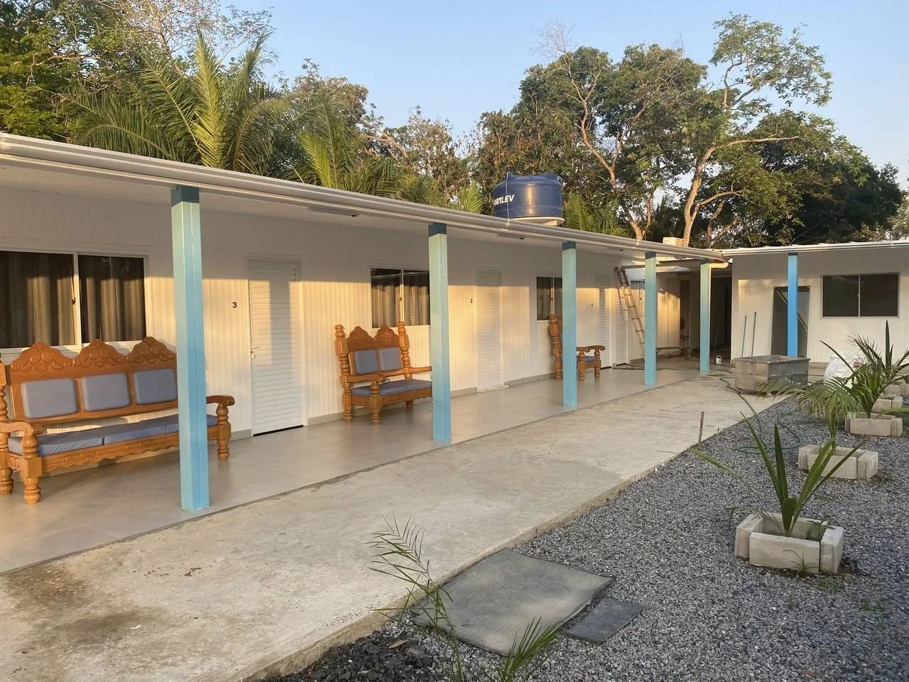 Covered walkway in front of the numbered rooms at Dona Onça Jaguar Lodge, with wooden benches, blue columns and palm trees