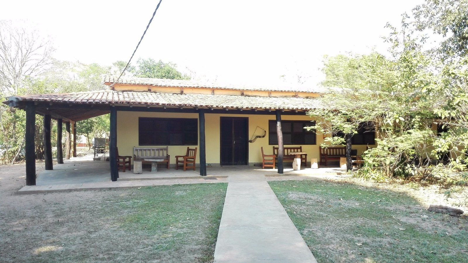 Main building of Pouso Alegre Lodge, a yellow single-storey house with a tiled roof, a wide veranda with wooden benches and chairs, and trees around a lawn