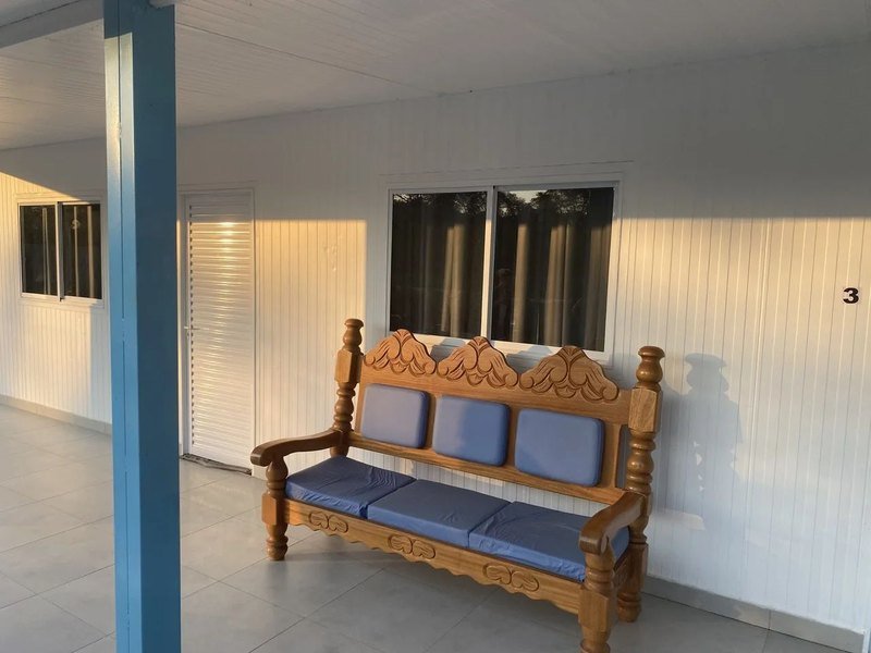 A carved wooden bench with blue cushions next to a room door and window, under the covered walkway