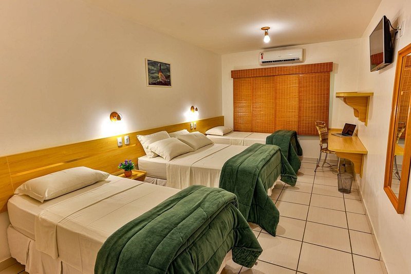 Apartment with three beds with green comforters, an air conditioner, a wall-mounted TV, a desk and a mirror