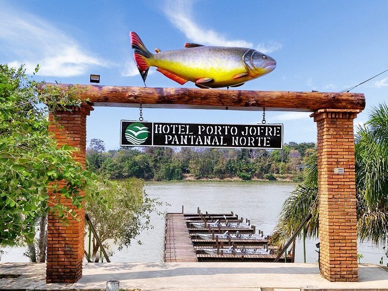 Entrance arch with a fish sculpture and the hotel sign, leading to a pier with boats on the river