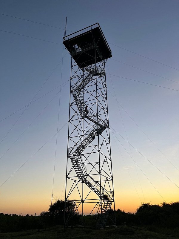 Tall steel observation tower with zigzag stairs and a covered platform, silhouetted against the dusk sky, with a person climbing the stairs
