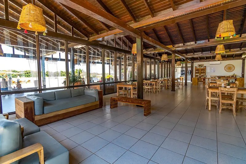 Restaurant with wooden tables and chairs, woven pendant lamps and large glass windows, with a lounge area in front