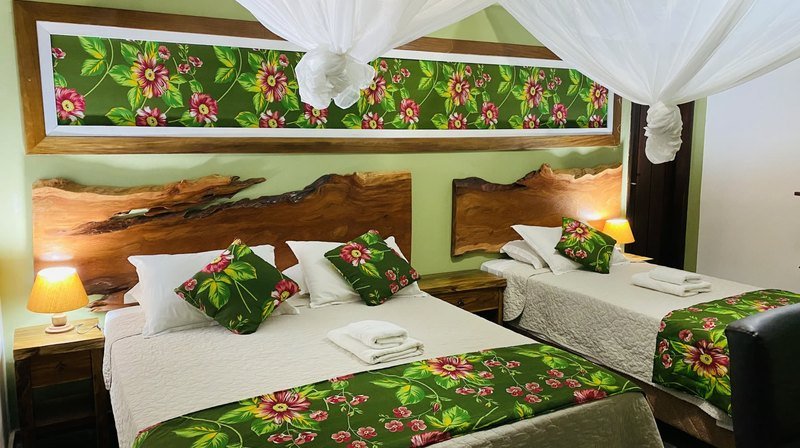 Room with a double bed and a single bed under mosquito nets, live-edge wooden headboards, green floral runners and cushions, and bedside lamps