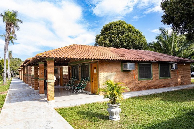 Single-storey brick building with a tiled roof, a covered veranda with chairs and air-conditioning units, beside a lawn and palm trees
